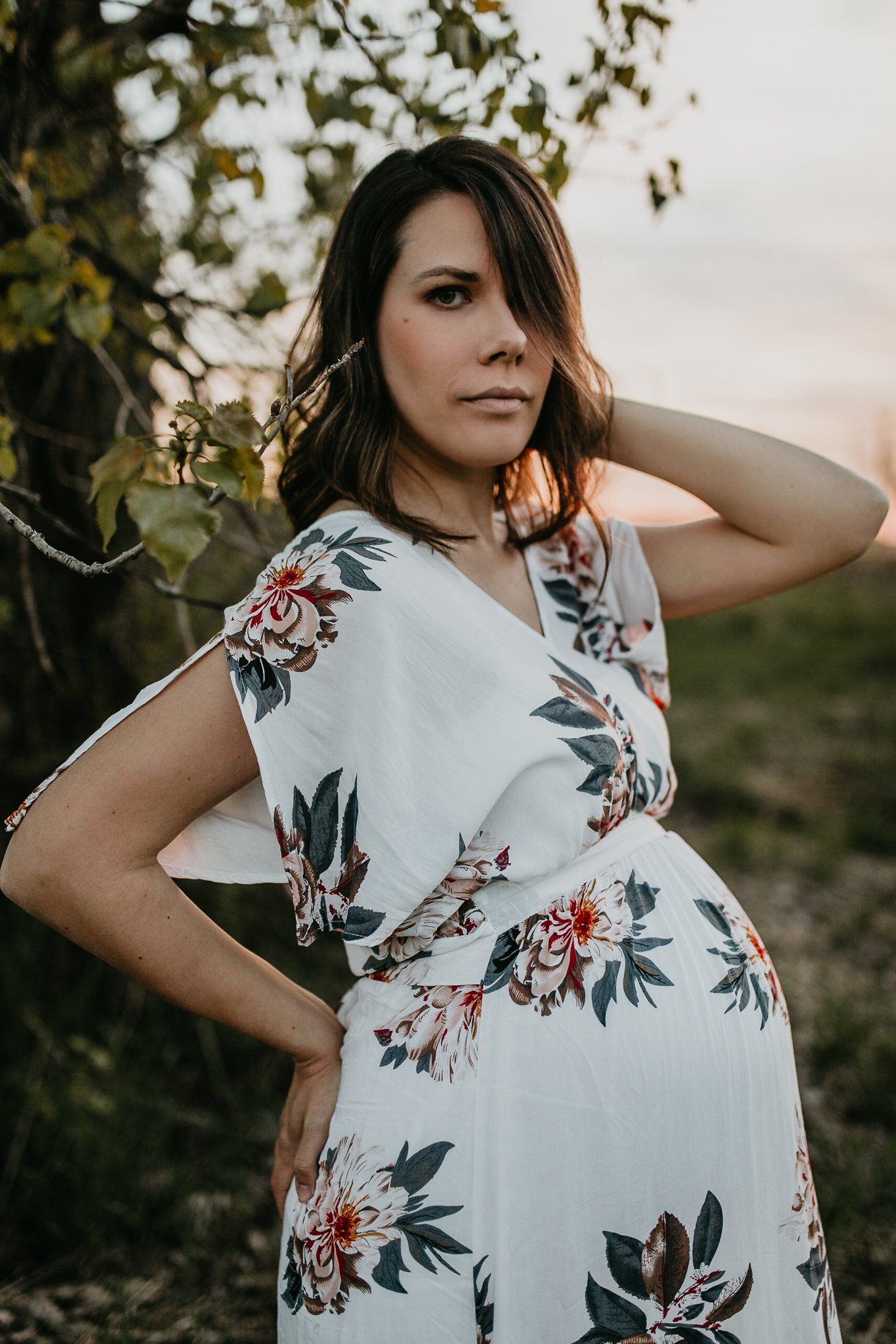 Expecting woman by a tree with one hand on her hip and one in her hair.
